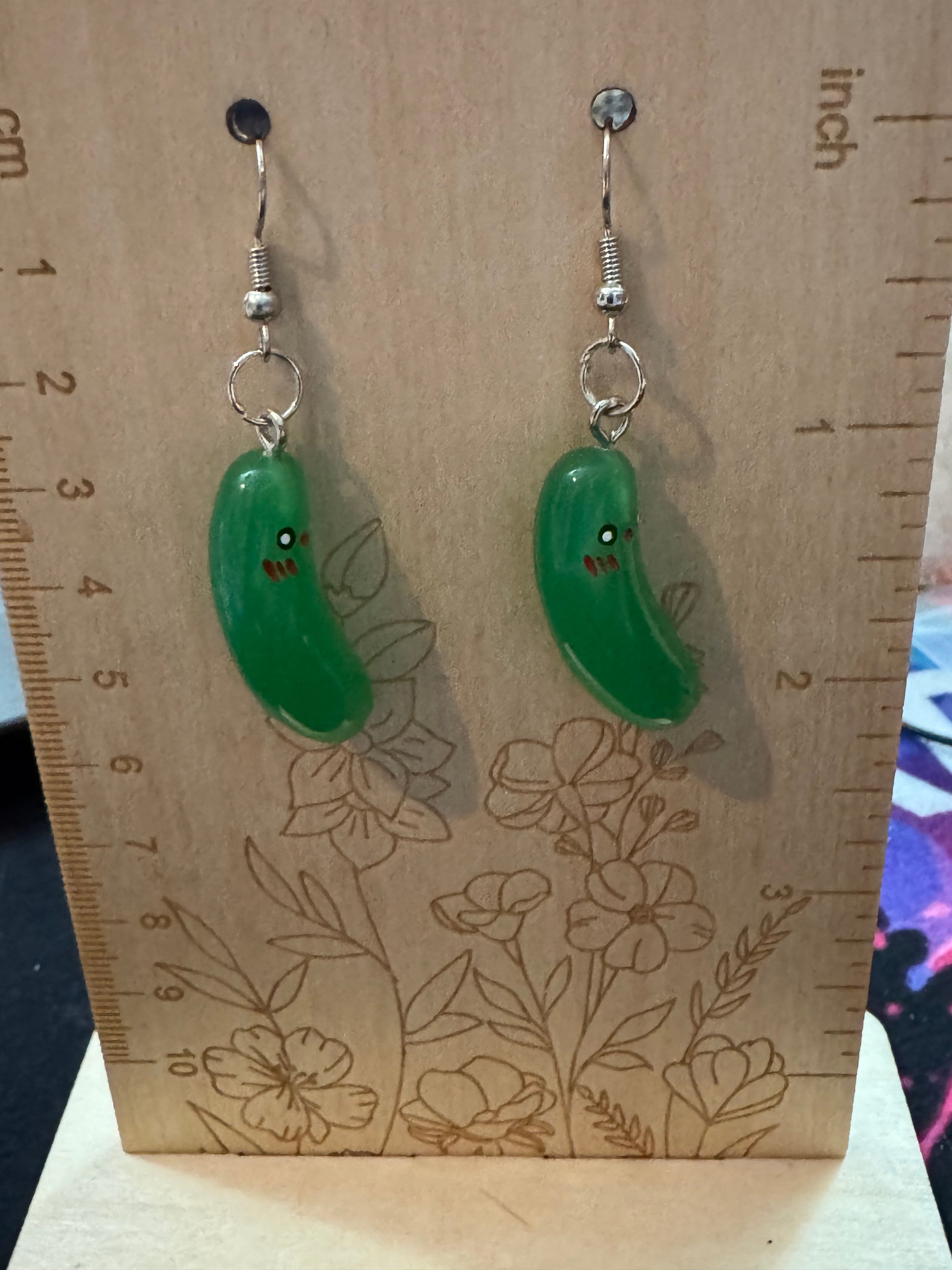 Two pickle earrings on a wooden board with floral patterns and a ruler for scale.