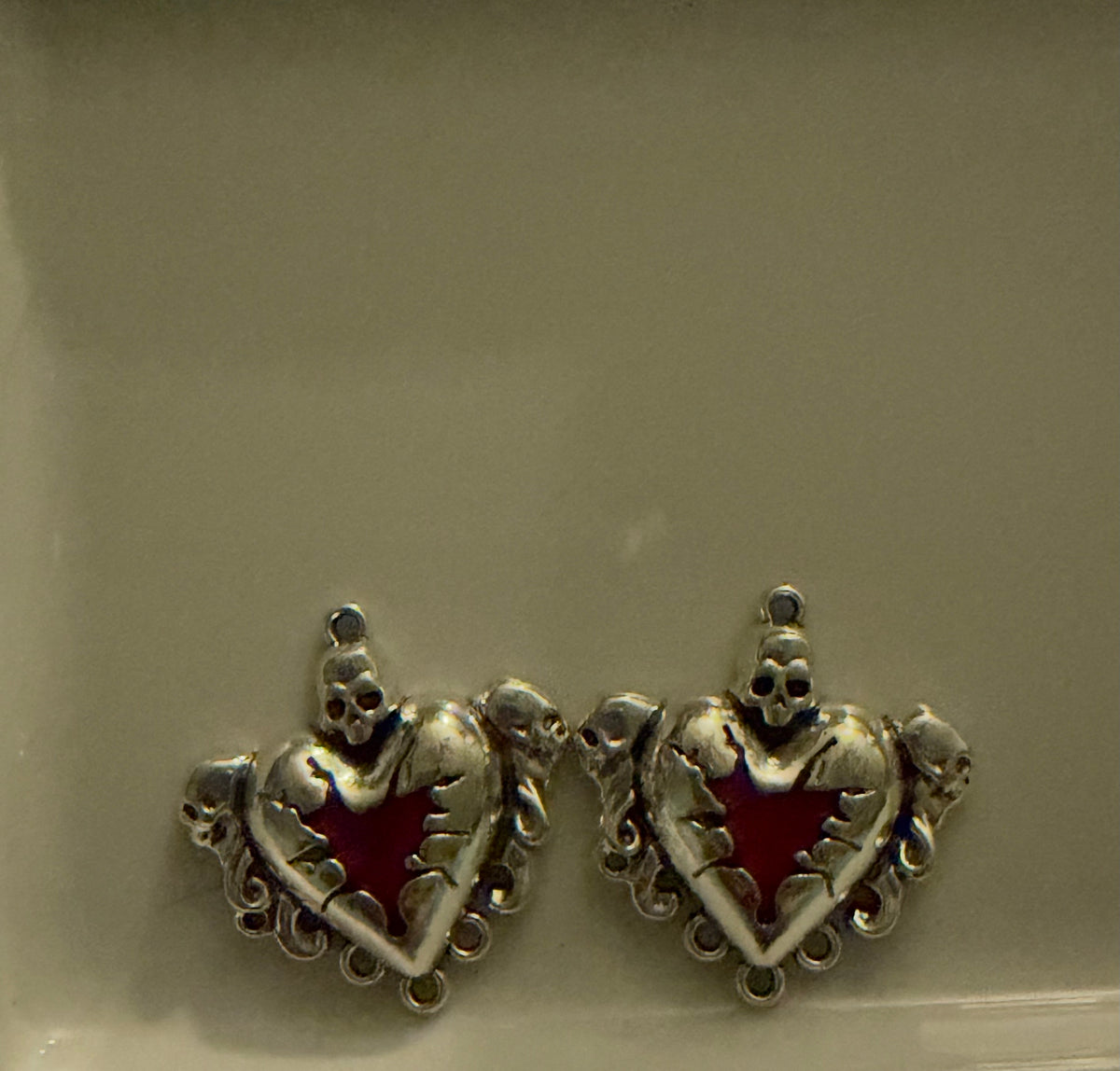 A pair of silver-toned gothic heart charms with skull details and a red enamel star center, displayed on a neutral beige surface.