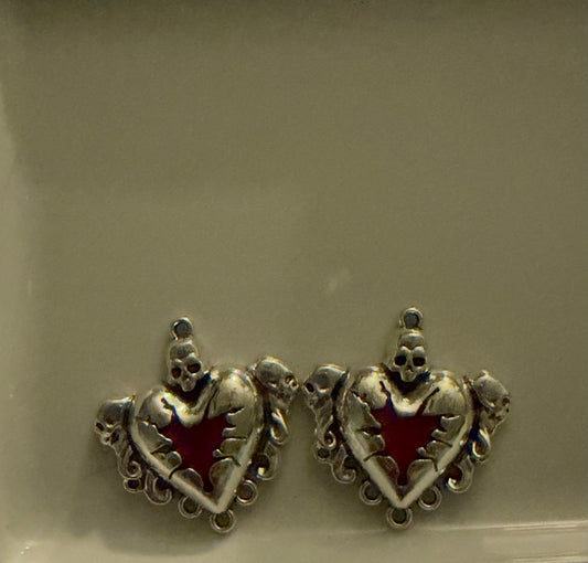A pair of silver-toned gothic heart charms with skull details and a red enamel star center, displayed on a neutral beige surface.