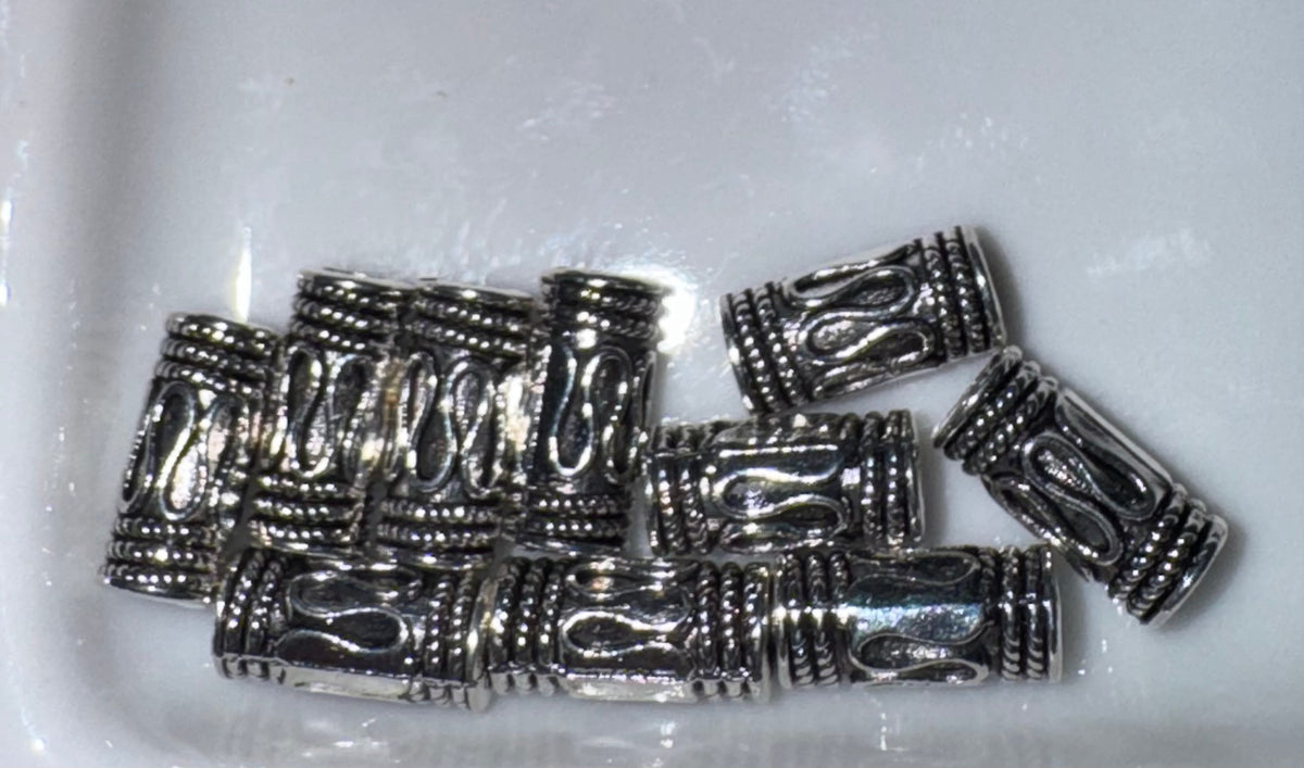 A group of small cylindrical silver-toned beads with textured scrollwork and ribbed ends, arranged on a white glossy surface.