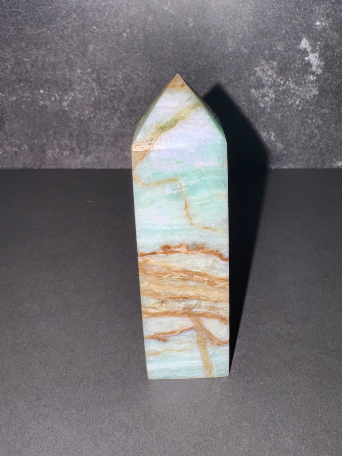 A polished crystal point tower showing natural green, blue, and tan marbling, standing upright on a dark gray surface against a textured gray background.