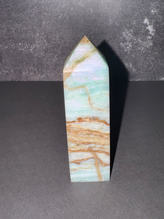 A polished crystal point tower showing natural green, blue, and tan marbling, standing upright on a dark gray surface against a textured gray background.