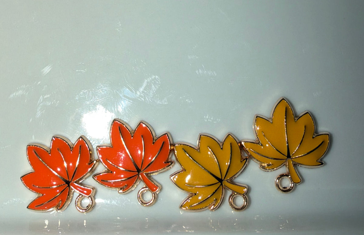 Four maple leaf-shaped charms with gold-tone metal edges and small hanging loops, shown in red-orange, orange, yellow, and mustard colors on a light gray background.