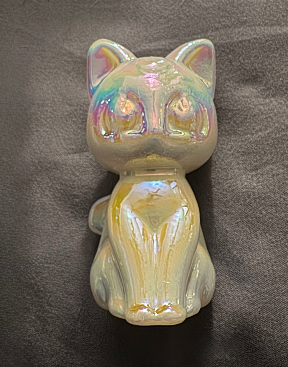 An iridescent stone cat figurine with a glossy, shifting pastel finish, sitting on a dark gray textured fabric background.