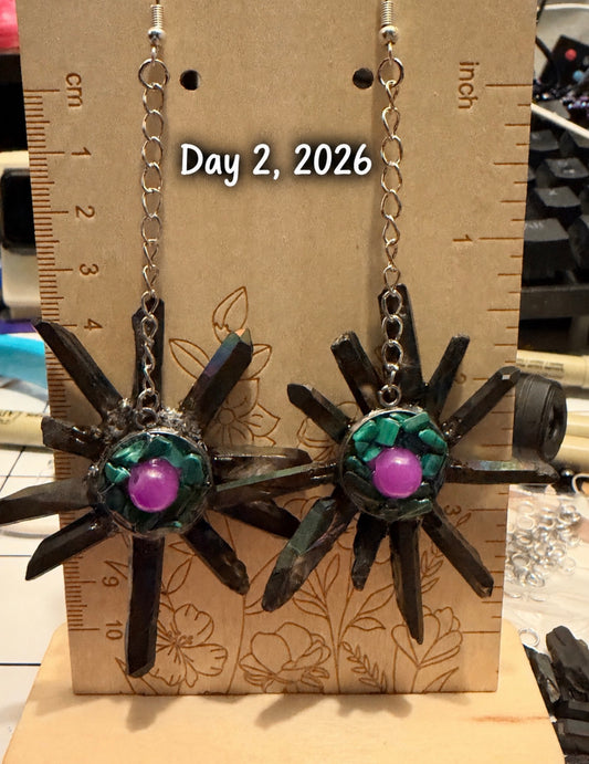 A pair of handmade spider earrings displayed on a cardboard card with inch rulers for scale. The earrings have dark brown spider bodies, green mosaic accents, and a shiny purple center bead, hanging from silver chains.