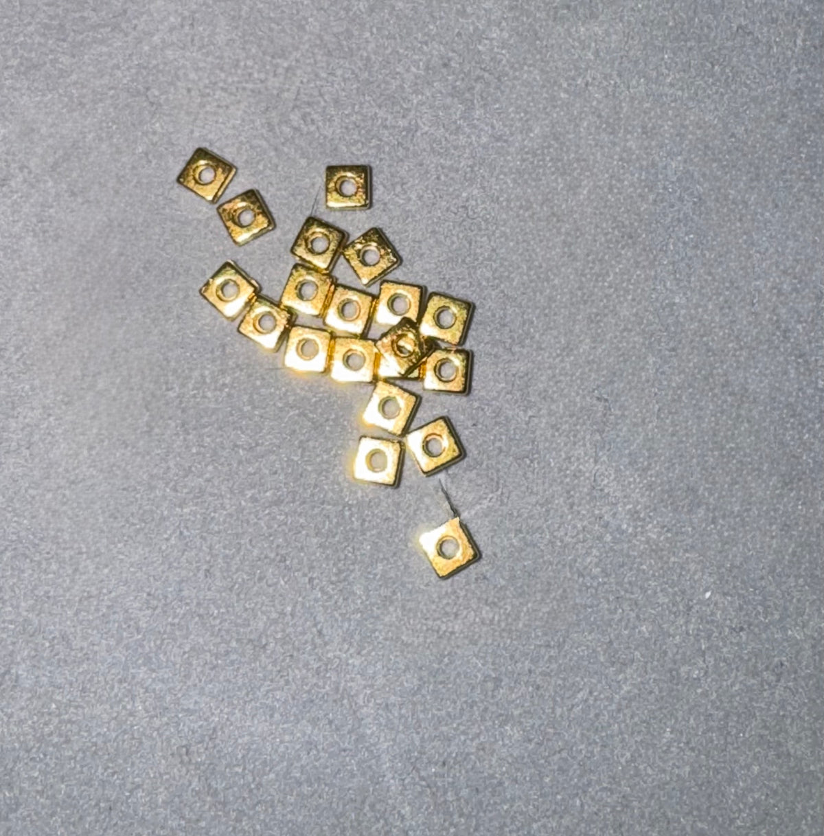 A cluster of small square gold metal components with circular cutouts, scattered on a textured gray surface.