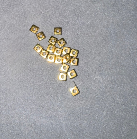 A cluster of small square gold metal components with circular cutouts, scattered on a textured gray surface.
