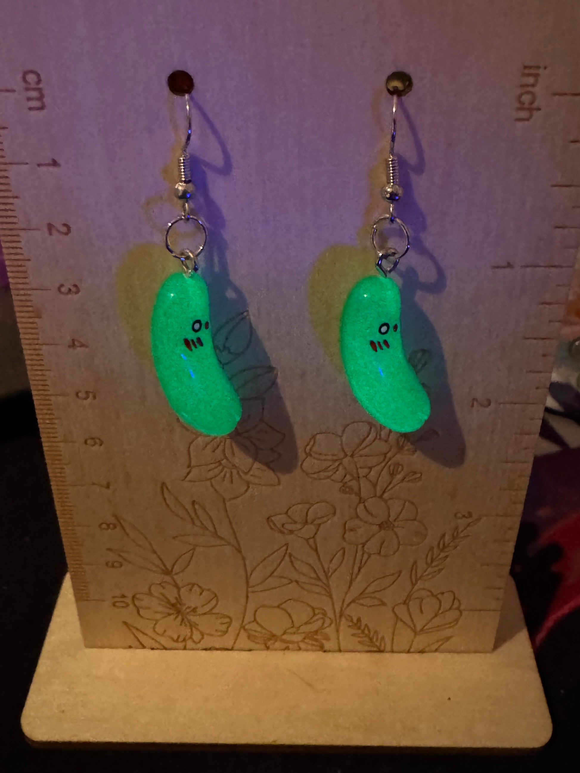 Two pickle earrings with face designs on a wooden board with a ruler.