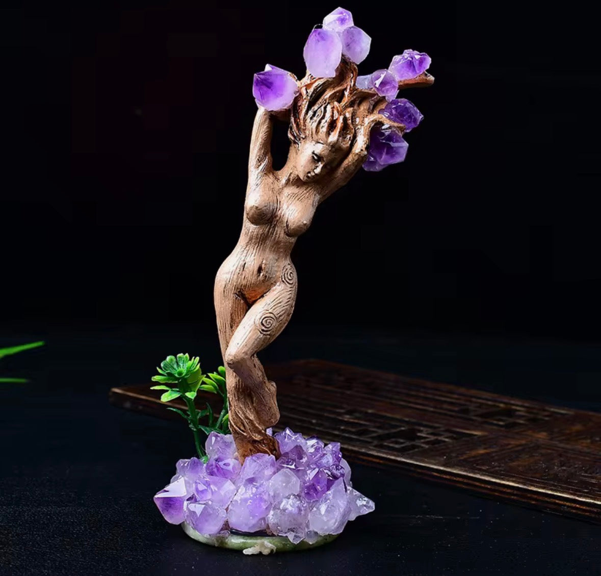 A decorative figurine of a nude female form made from natural wood, holding purple crystal flowers above her head, mounted on a cluster of purple crystals with a small green plant accent, set against a dark background.