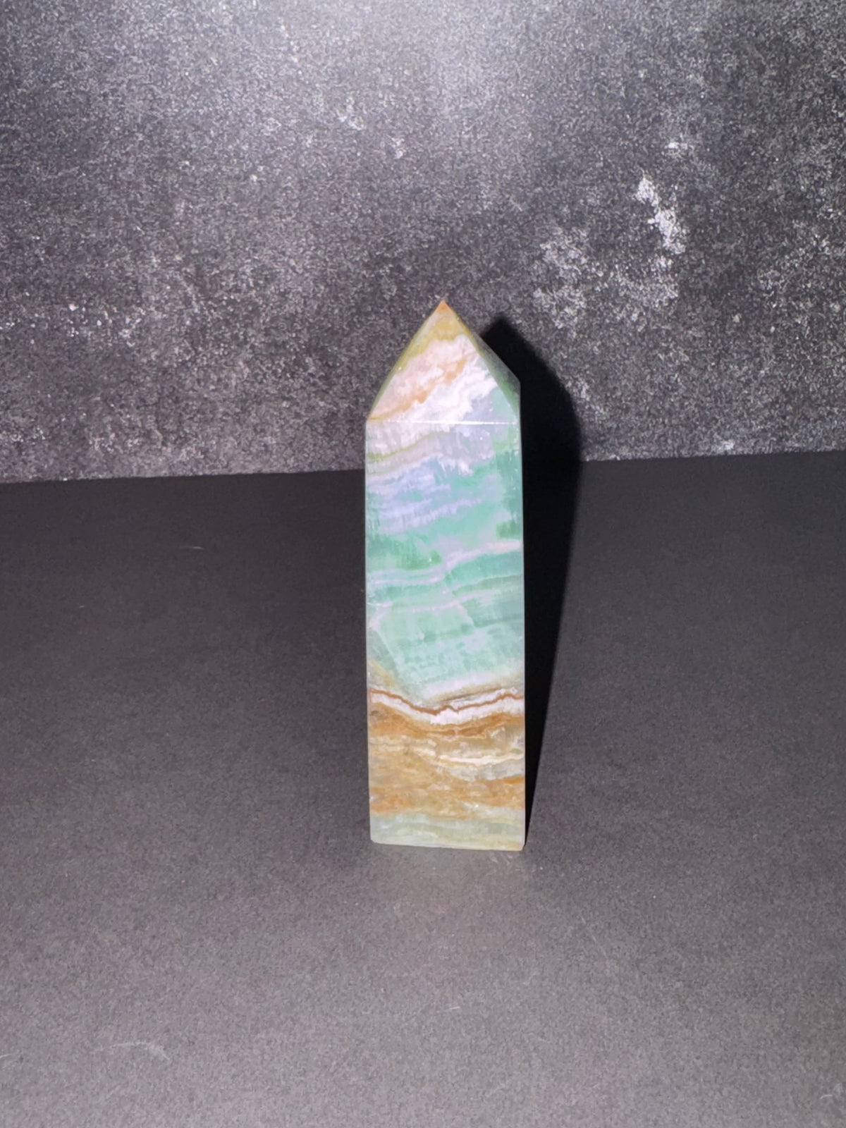 A polished natural crystal point, featuring bands of light green, white, and tan, standing upright on a dark gray surface against a textured dark gray background.