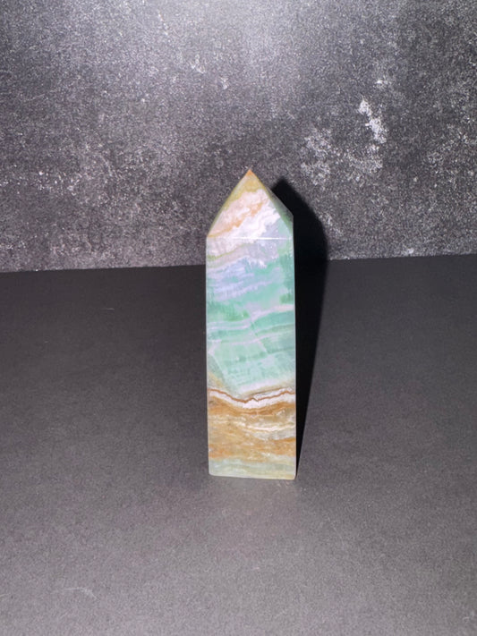 A polished natural crystal point, featuring bands of light green, white, and tan, standing upright on a dark gray surface against a textured dark gray background.