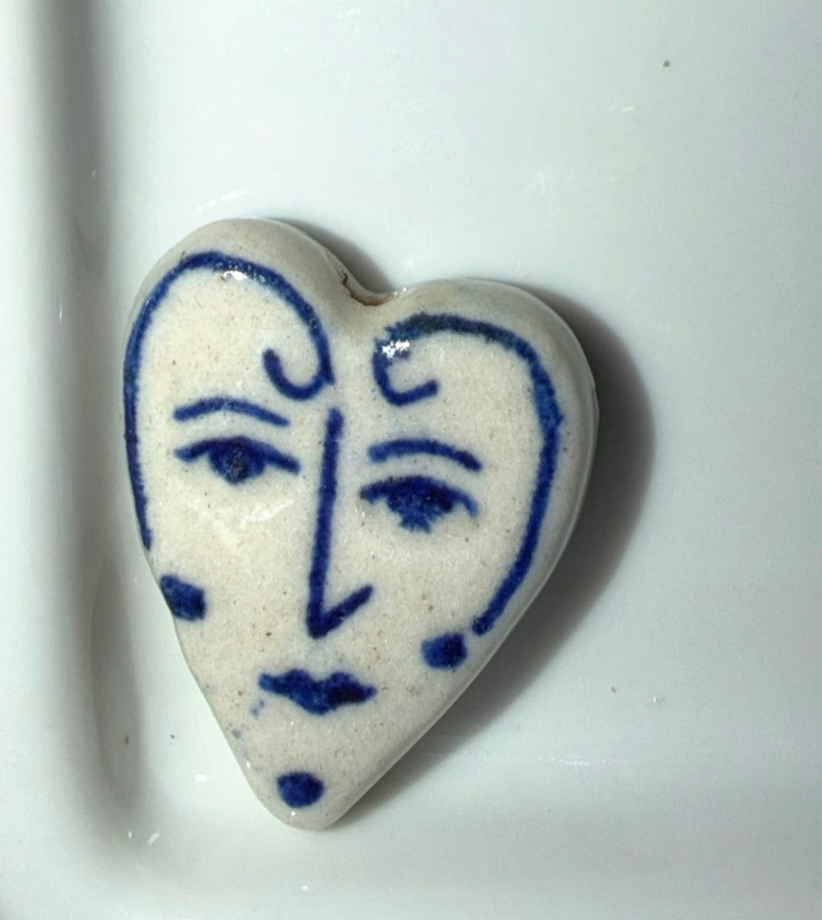 A heart-shaped ceramic charm with a stylized blue face design on a white background, resting on a white surface.