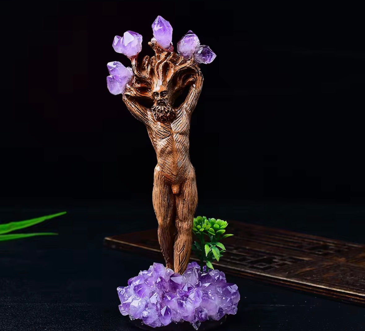A carved wooden figurine of a nude human form with arms raised, holding purple amethyst crystal points like flowers, mounted on a cluster of purple crystals with a small green plant accent, set against a dark background.