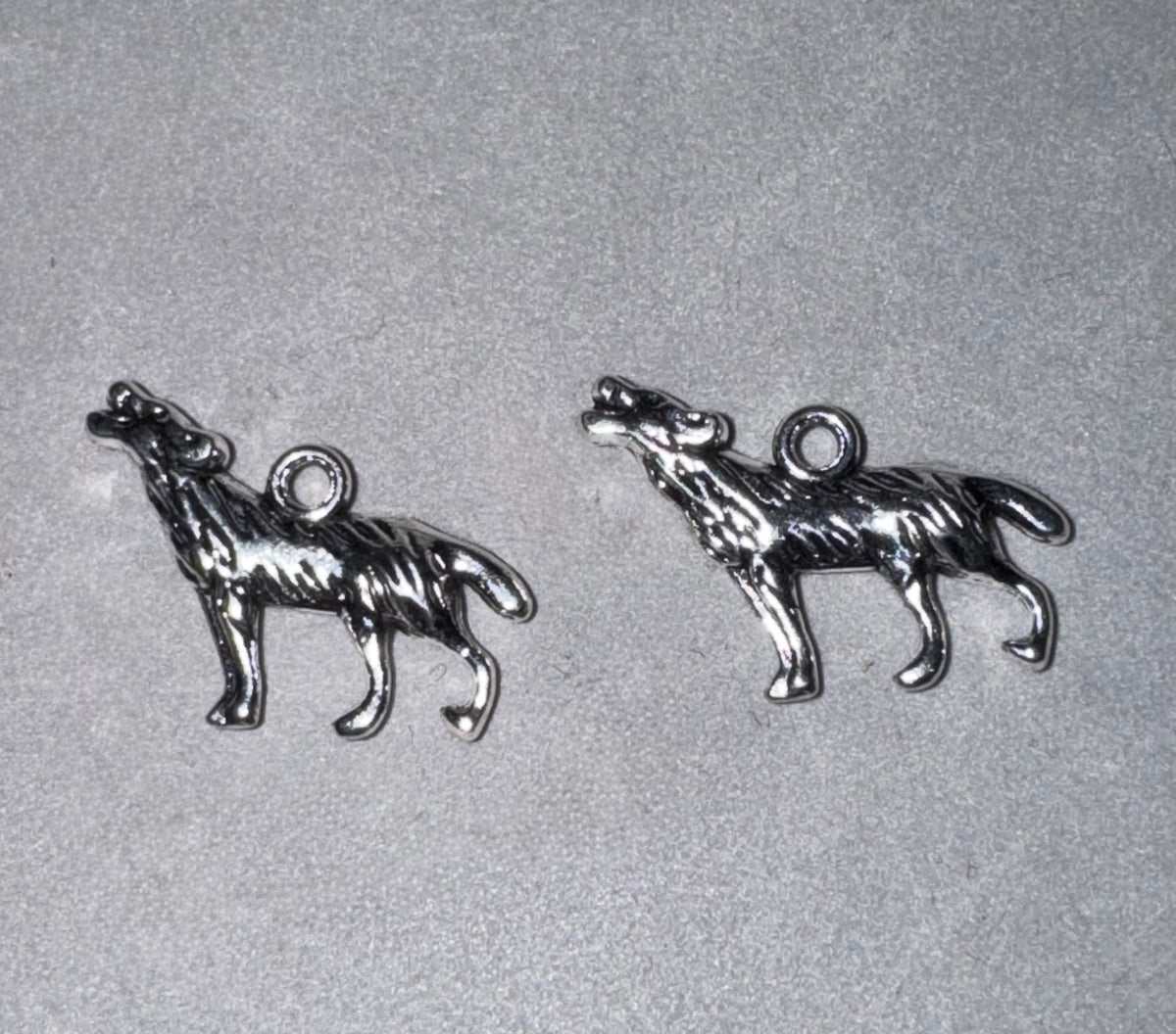Two small silver-toned metal charms shaped like howling wolves, shown on a light gray textured background.