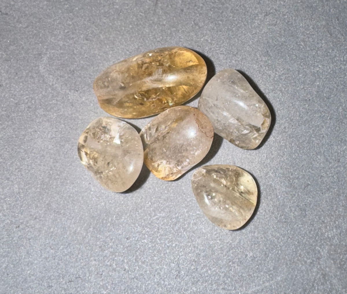 Five polished tumbled citrine gemstones in varying shapes and translucent colors, including yellow, clear, and smoky tones, arranged on a textured gray background.