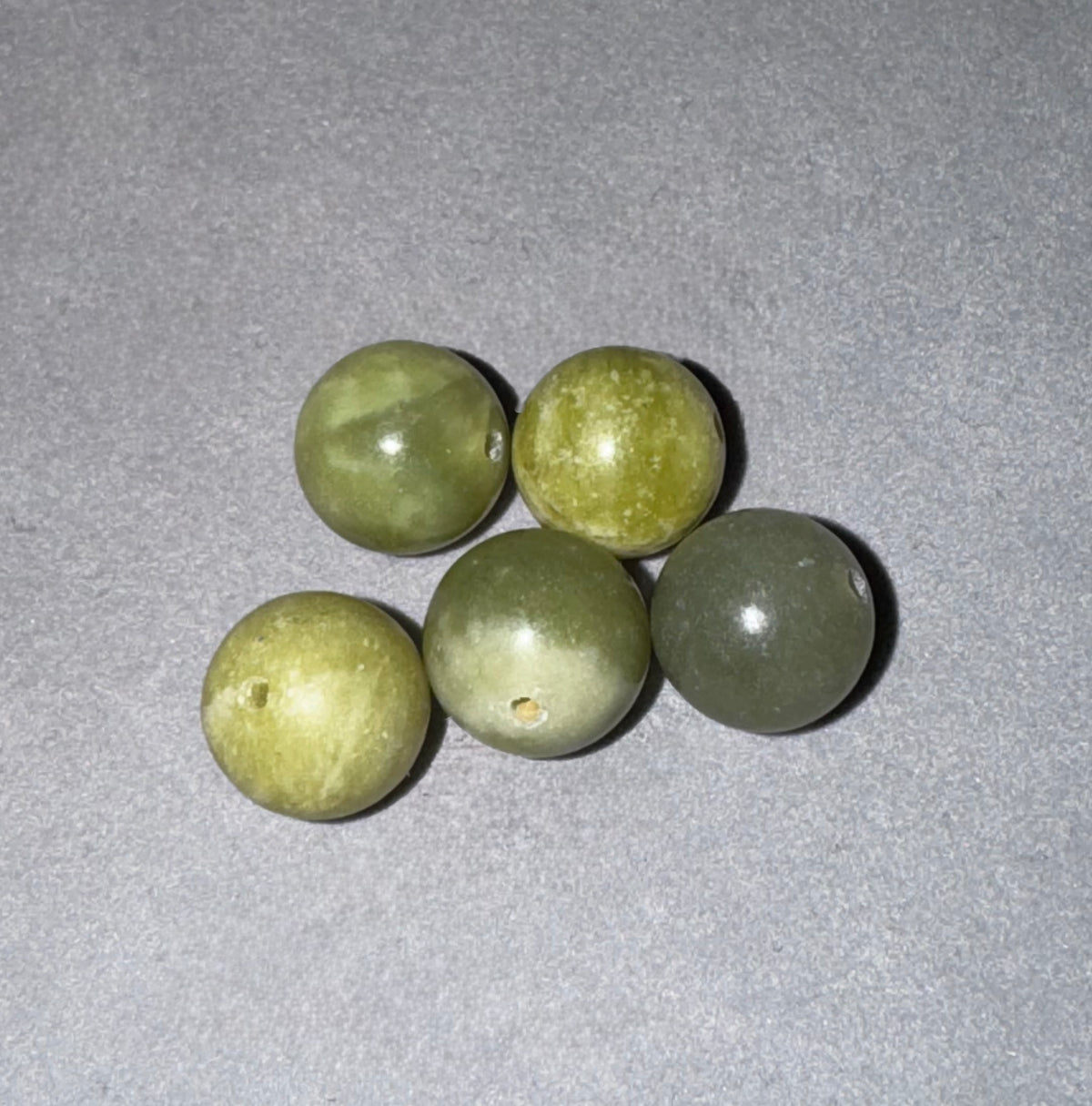 Five round, polished stone beads in shades of olive green and yellow-green, arranged on a textured gray background.