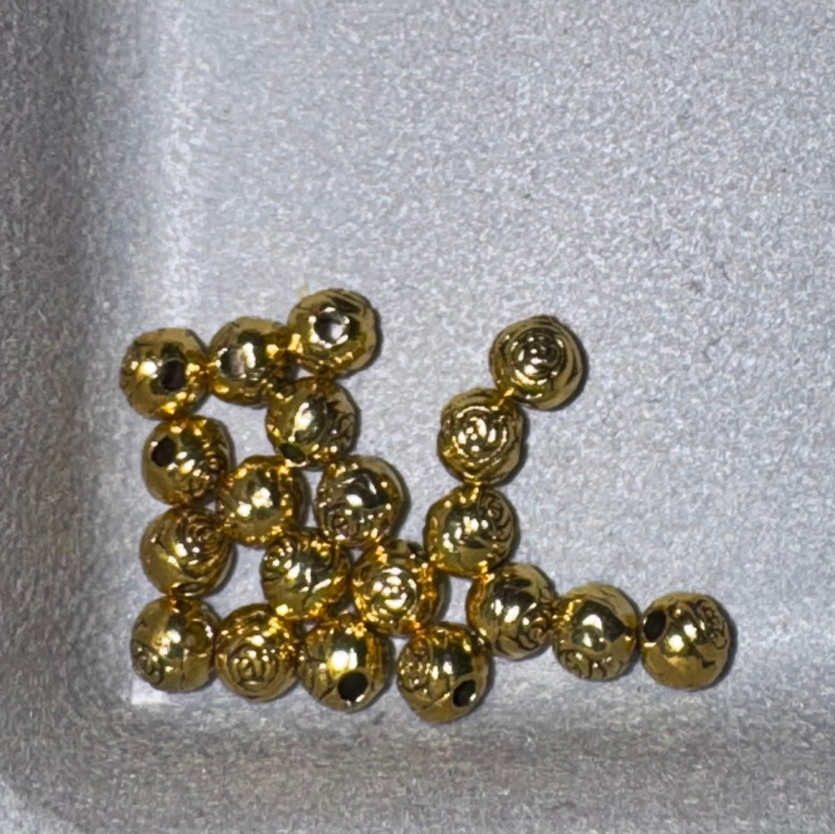 A group of small, shiny gold metallic beads with a textured, hammered finish, arranged on a light gray textured foam surface.