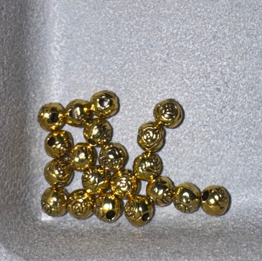 A group of small, shiny gold metallic beads with a textured, hammered finish, arranged on a light gray textured foam surface.