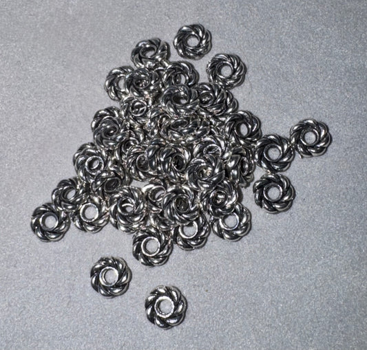 A pile of small metallic silver chain links with a twisted or swirled design, scattered on a textured gray background.