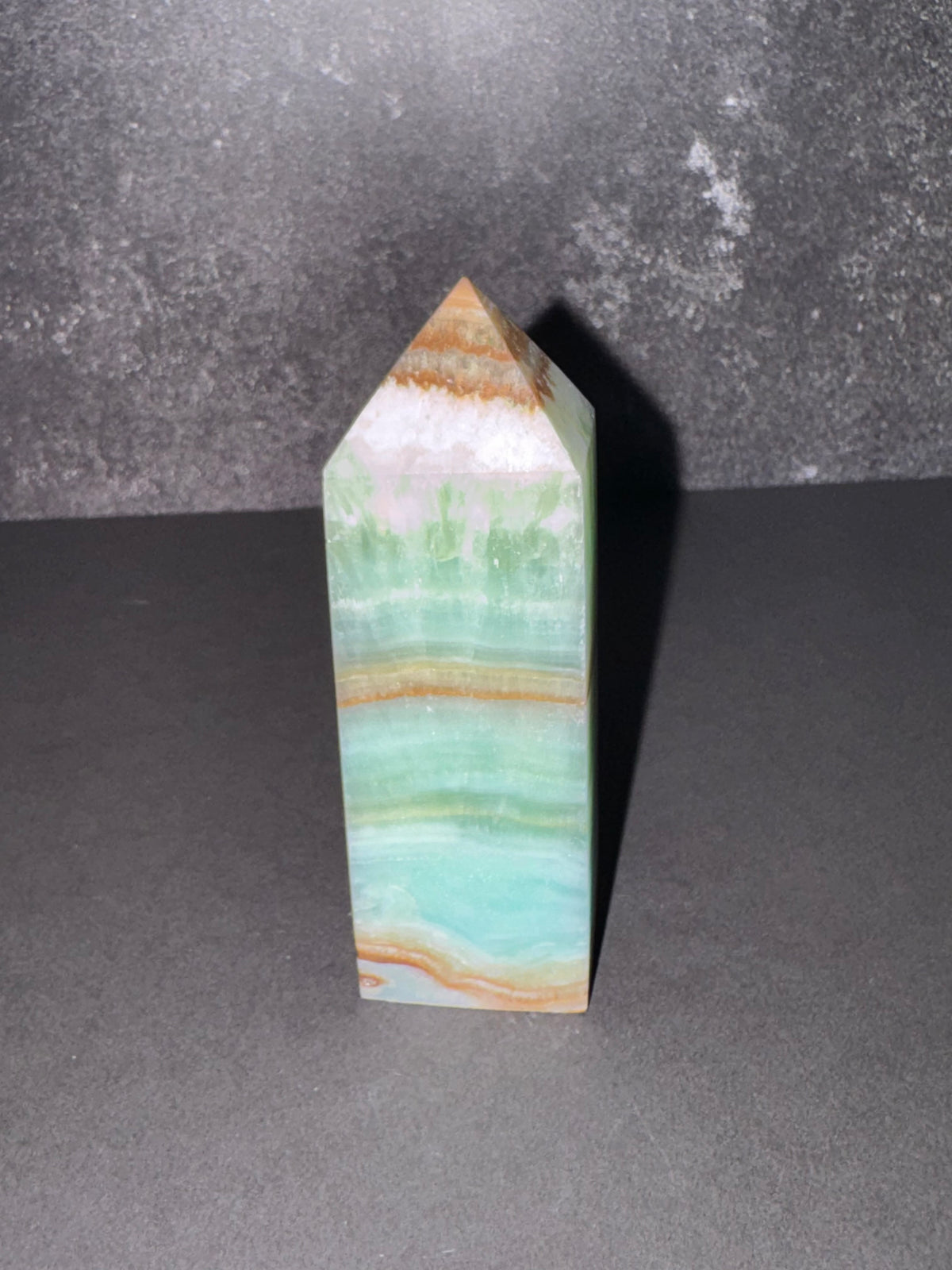 A polished natural crystal point, featuring horizontal bands of light green, turquoise, and tan, standing upright on a dark gray surface against a textured dark gray background.