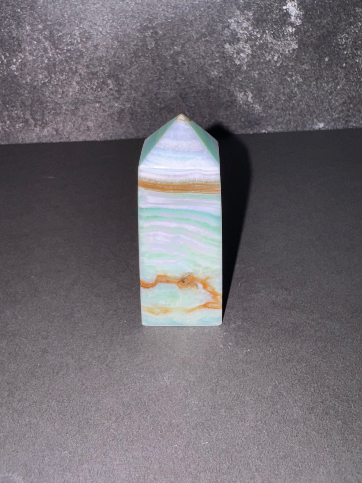 A polished natural stone pyramid with wavy bands of light green, white, and tan, standing upright on a dark gray surface against a textured gray background.