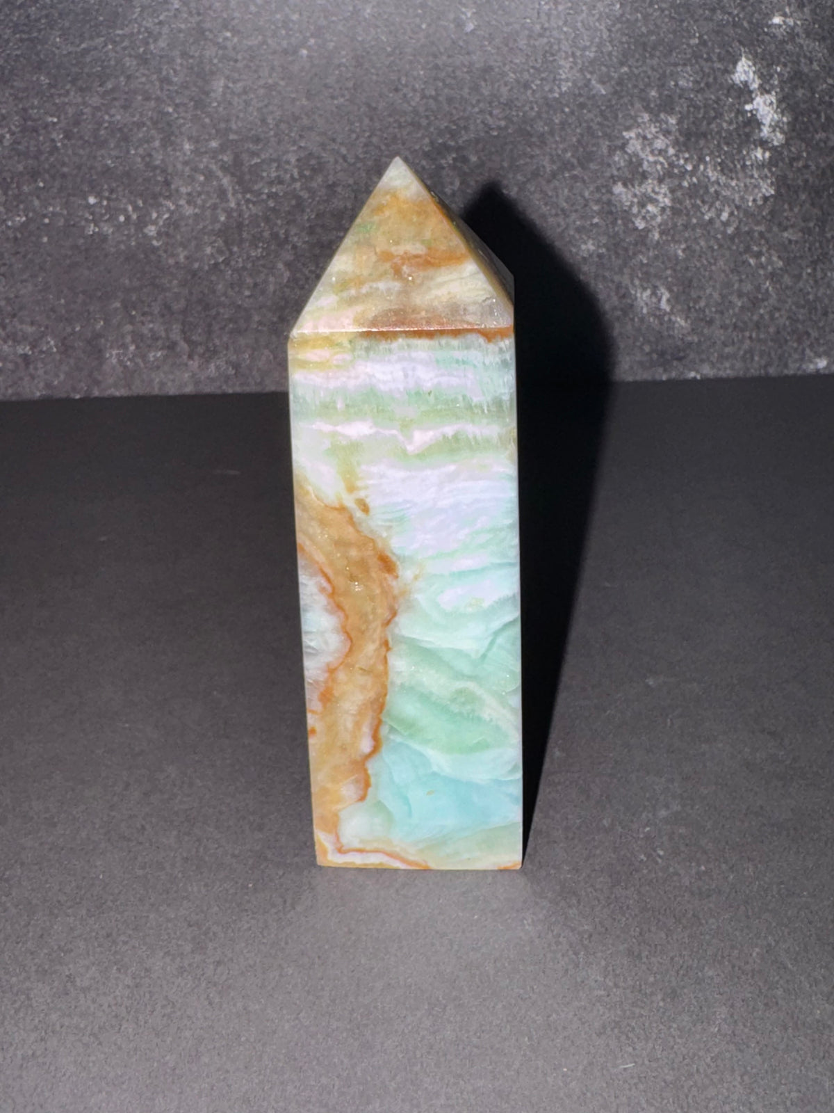 A polished natural stone pyramid tower, featuring wavy bands of light green, white, and tan, standing on a dark gray surface against a textured dark gray background.