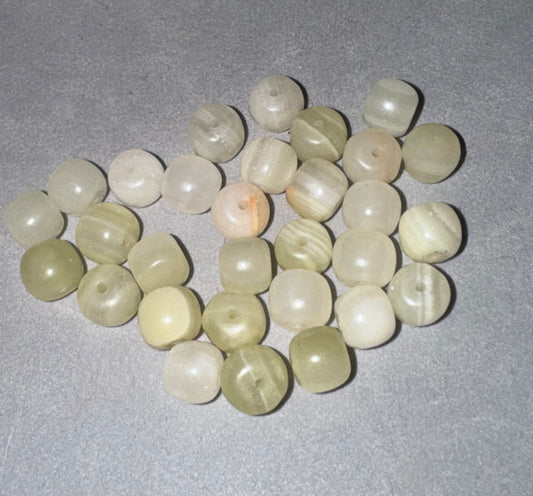 A group of small, round, polished stone beads in various light shades of beige, cream, and pale green, arranged on a textured gray surface.