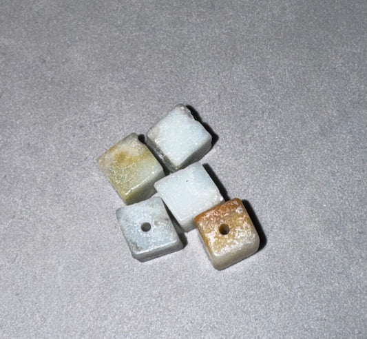 Four small square stone beads with holes, featuring natural marbled textures in white, light gray, and light brown, arranged on a textured gray background.