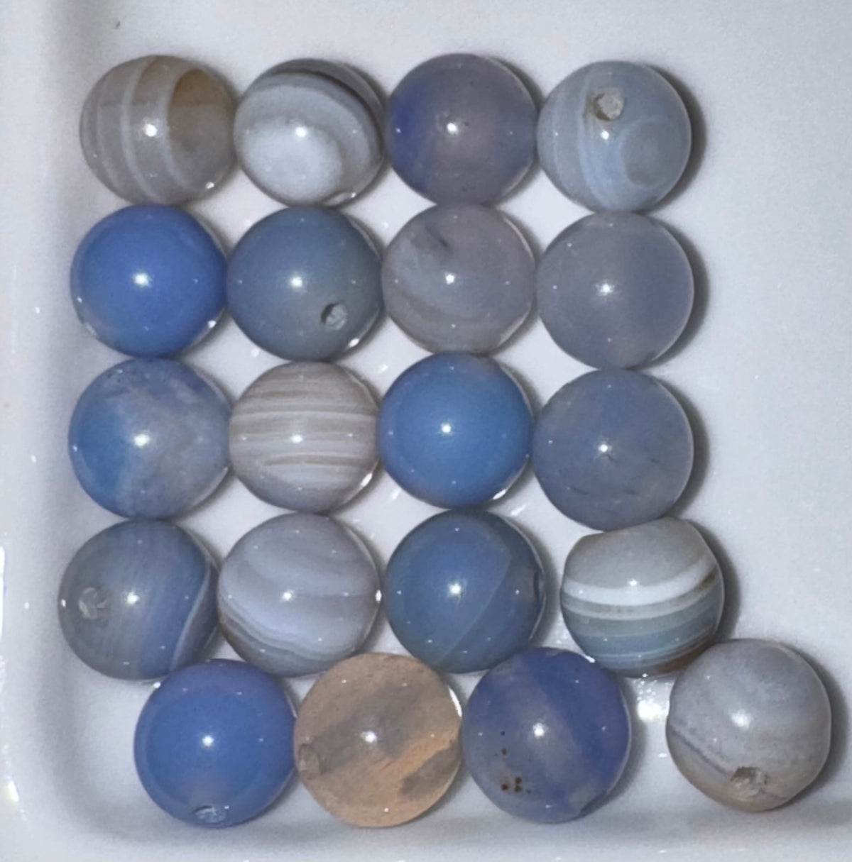 A set of round polished gemstone beads arranged in rows on a white surface. The beads are primarily shades of blue, gray, and white, with some showing banded agate-like patterns and one translucent tan bead.