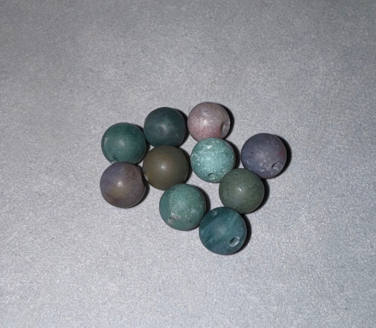 Nine round, matte stone beads in various earthy colors including green, blue, purple, and brown, arranged on a textured gray background.