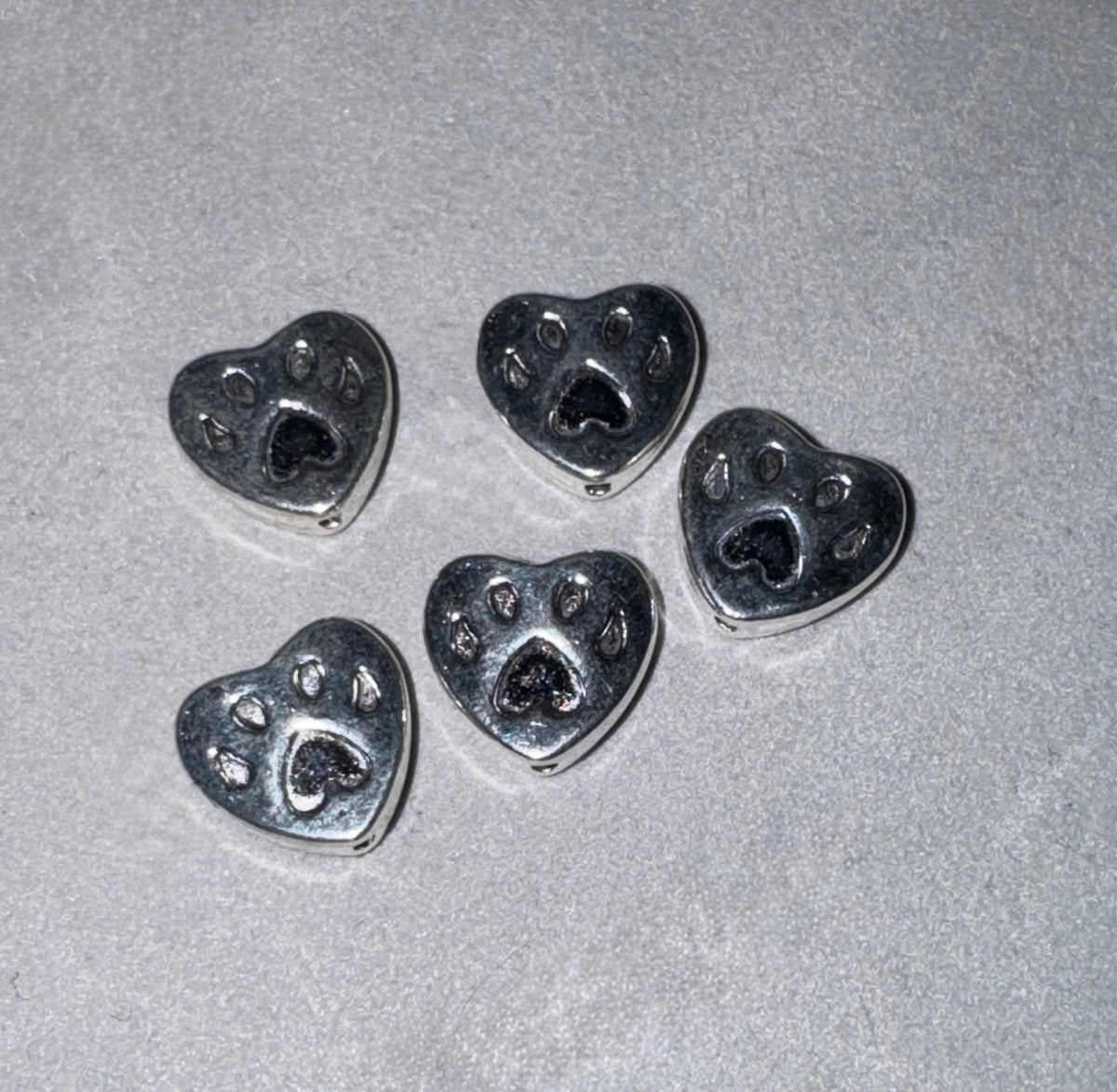 Five silver heart-shaped charms with paw print details, arranged on a textured light gray background.