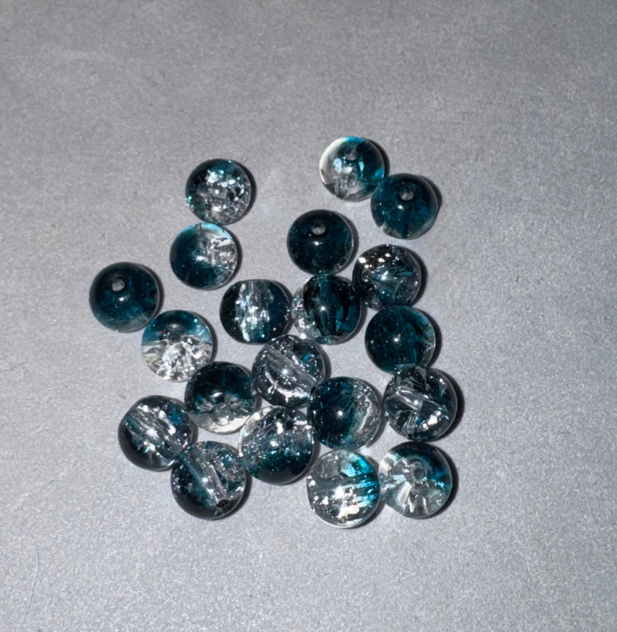 A group of round, dark teal glass beads with white glitter swirls, arranged on a textured gray background.