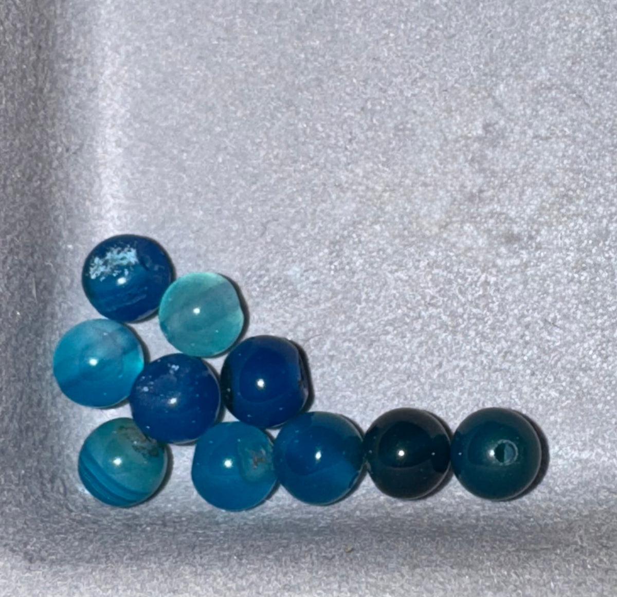 A group of round blue beads in various shades of blue, teal, and dark navy, arranged on a textured light gray foam surface.