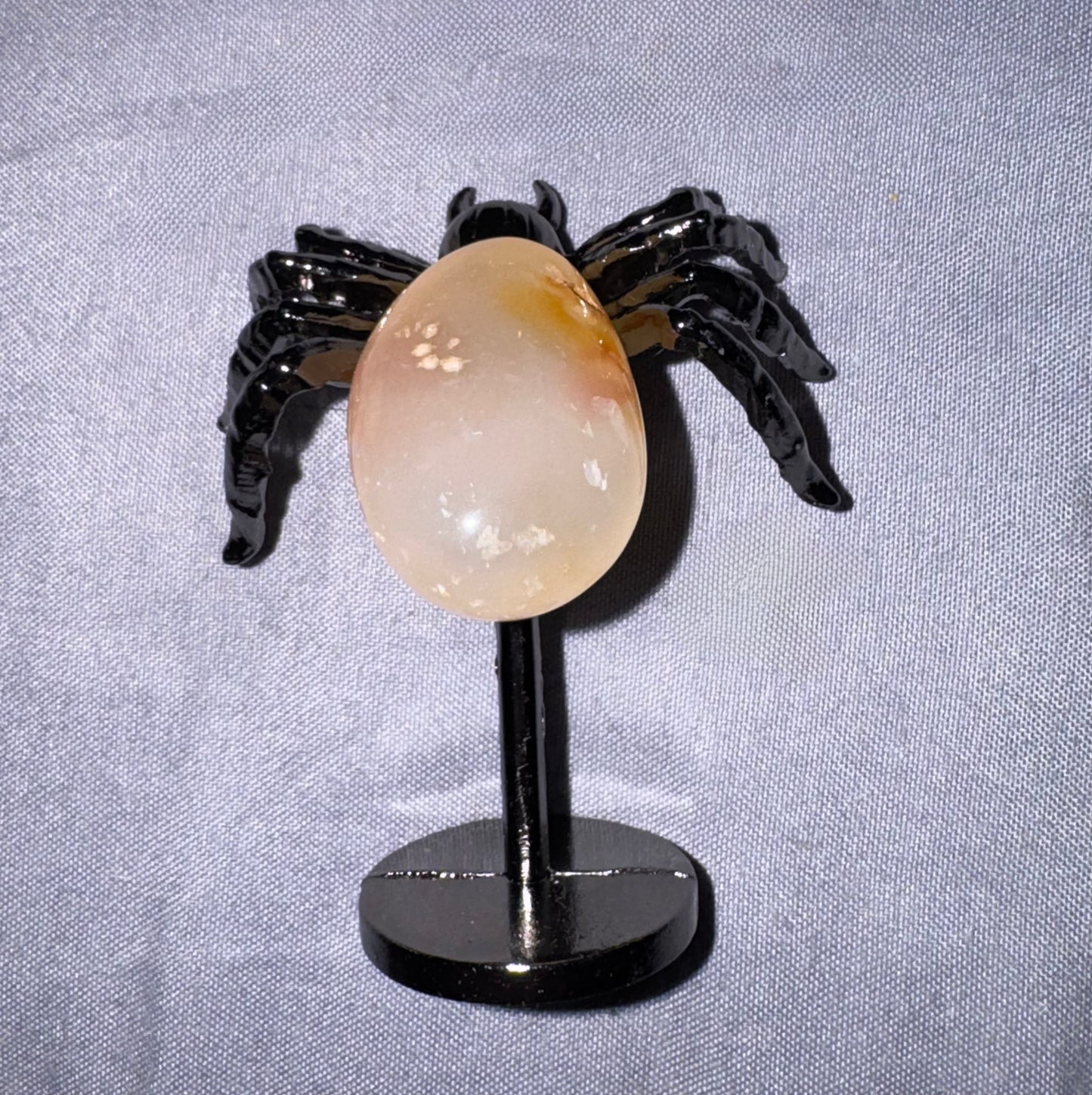 A polished peach-colored agate stone egg displayed on a black spider-shaped stand against a textured light gray fabric background.