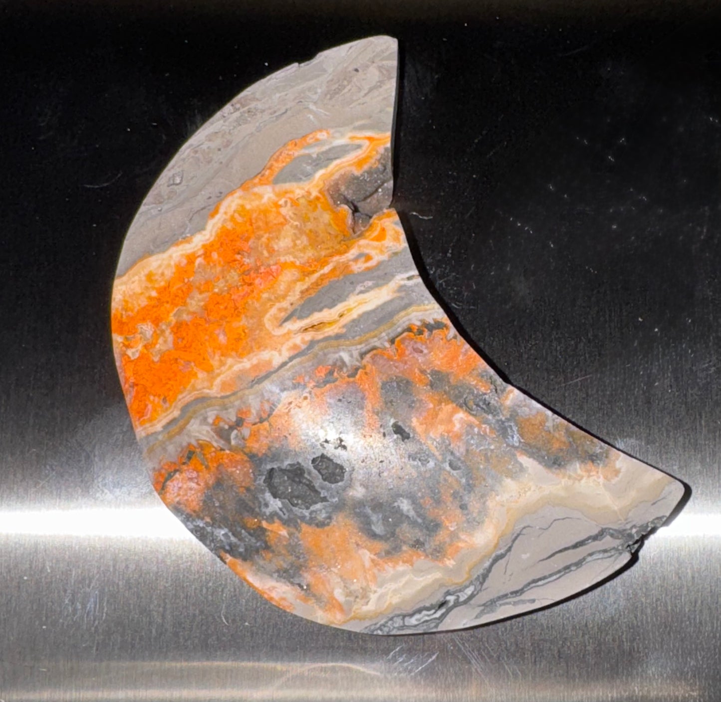 A crescent-shaped decorative stone plaque featuring vibrant orange, gray, and beige agate-like patterns, displayed on a brushed silver metal surface.