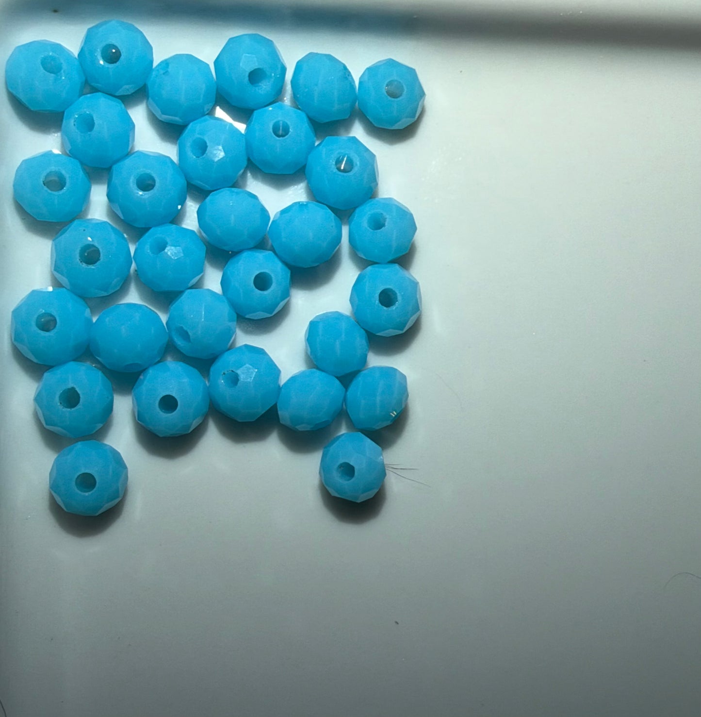 A pile of light blue faceted round beads with central holes, arranged on a plain light gray surface.