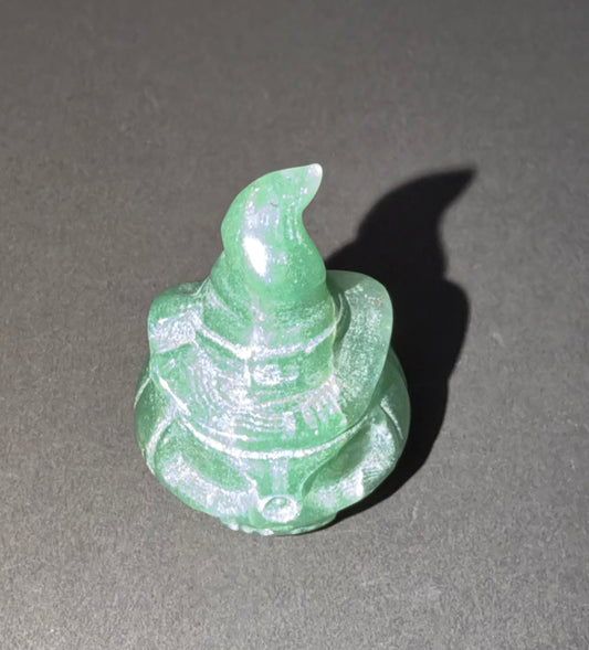 A small carved green stone figurine with a pointed top and textured body, photographed on a dark gray surface with a strong shadow to the right.