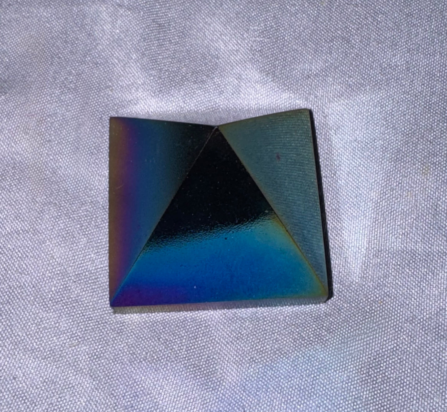 A square pyramid-shaped crystal with an iridescent finish, showing a color-shifting rainbow sheen of blue, green, and purple, resting on a textured white fabric background.