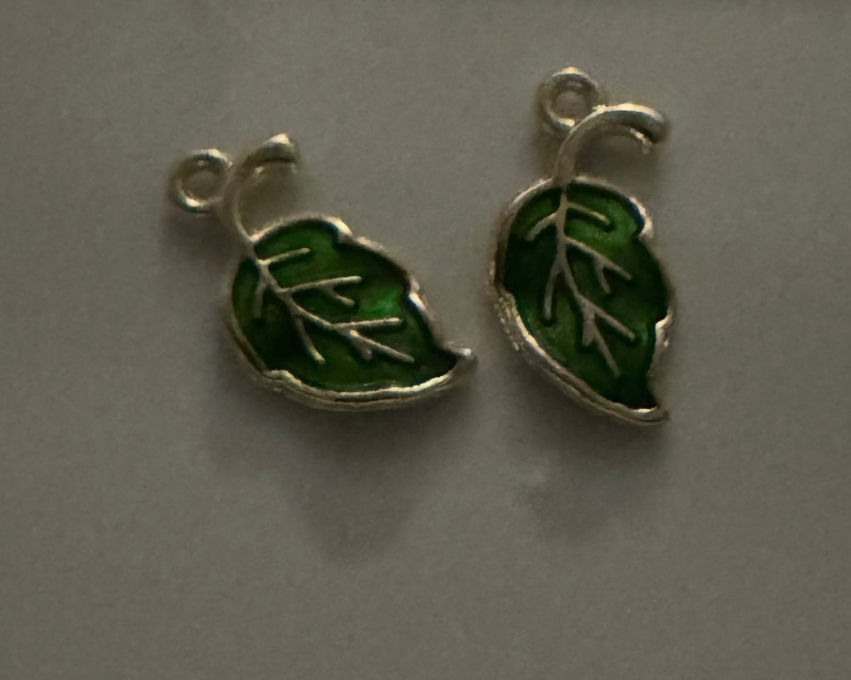 Two small silver leaf-shaped charms with green enamel fill, shown on a plain gray surface.
