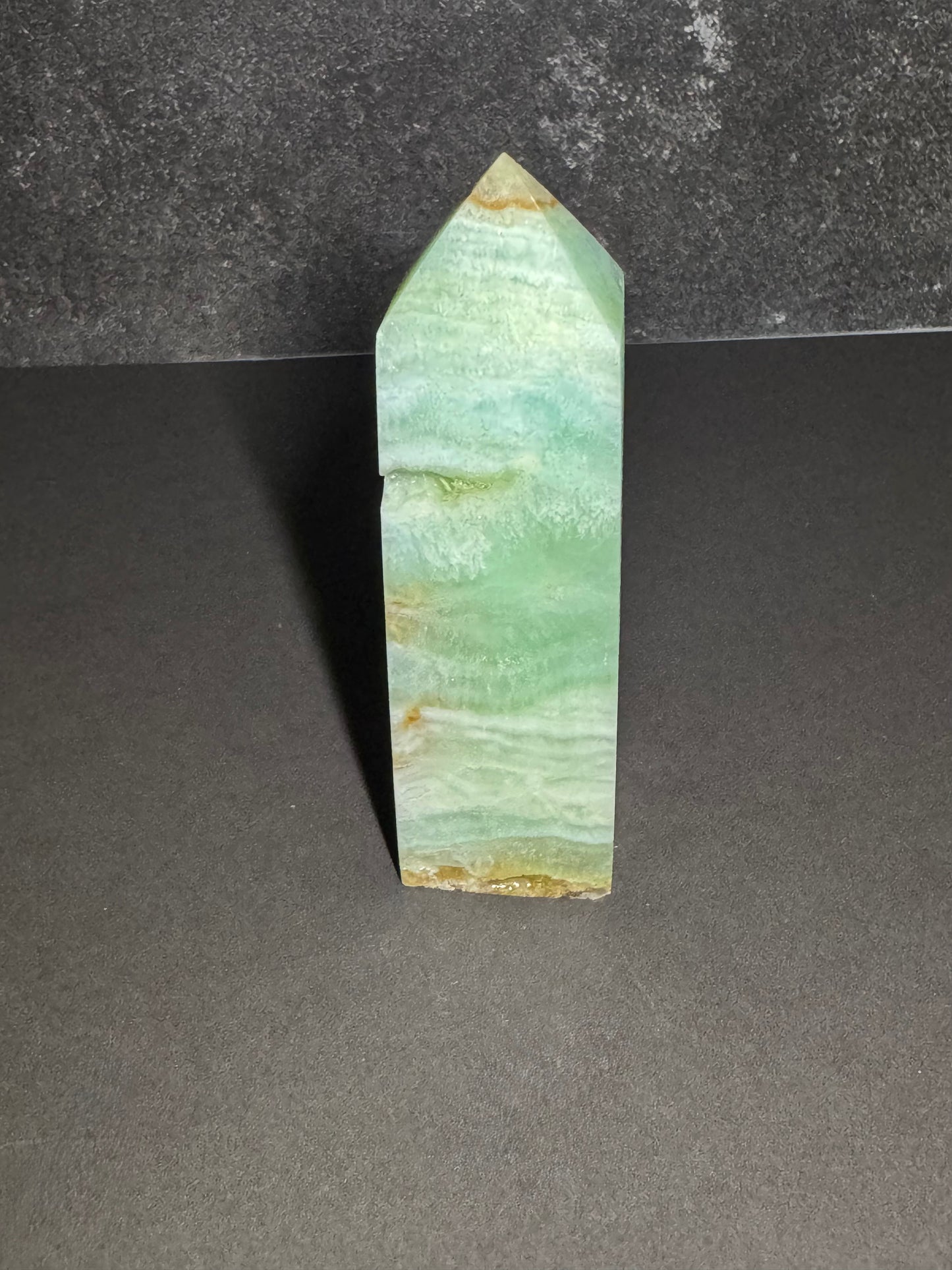 A polished natural crystal point, featuring light green and white banded patterns with small golden-brown mineral inclusions near the tip and base, standing upright on a dark gray surface.