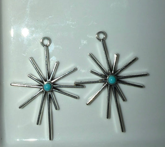 A pair of silver starburst dangle earrings with a small round turquoise stone in the center, displayed on a glossy white surface.