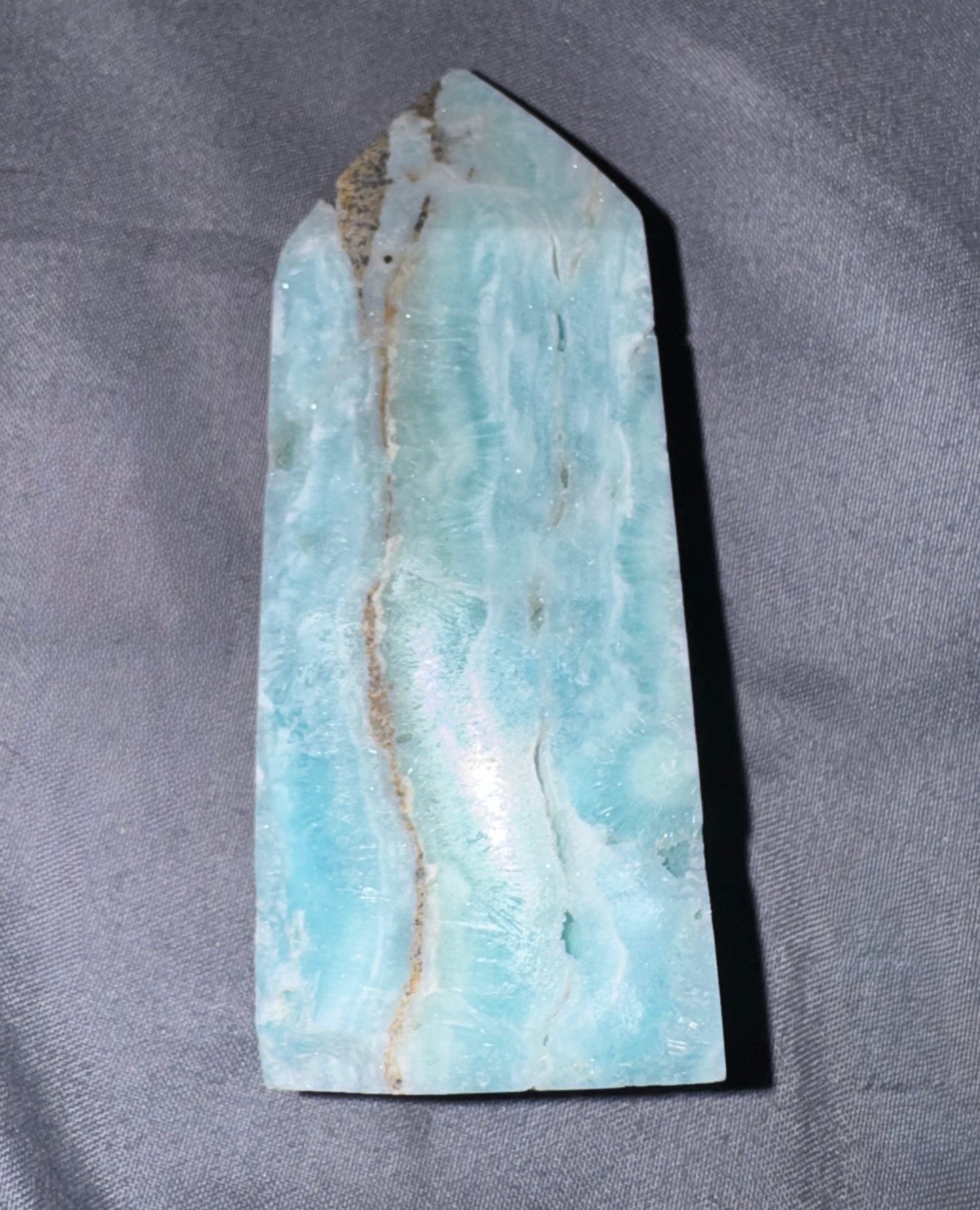 A polished natural stone or crystal with a light blue to turquoise coloration and brown veining, photographed upright on a gray fabric background.