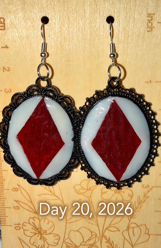 A pair of dangling earrings with oval silver-toned filigree frames, featuring white circular inserts with a red diamond in the center, hanging on a beige patterned surface with a ruler visible in the background and the text 'Day 20, 2026' overlaid at the bottom.