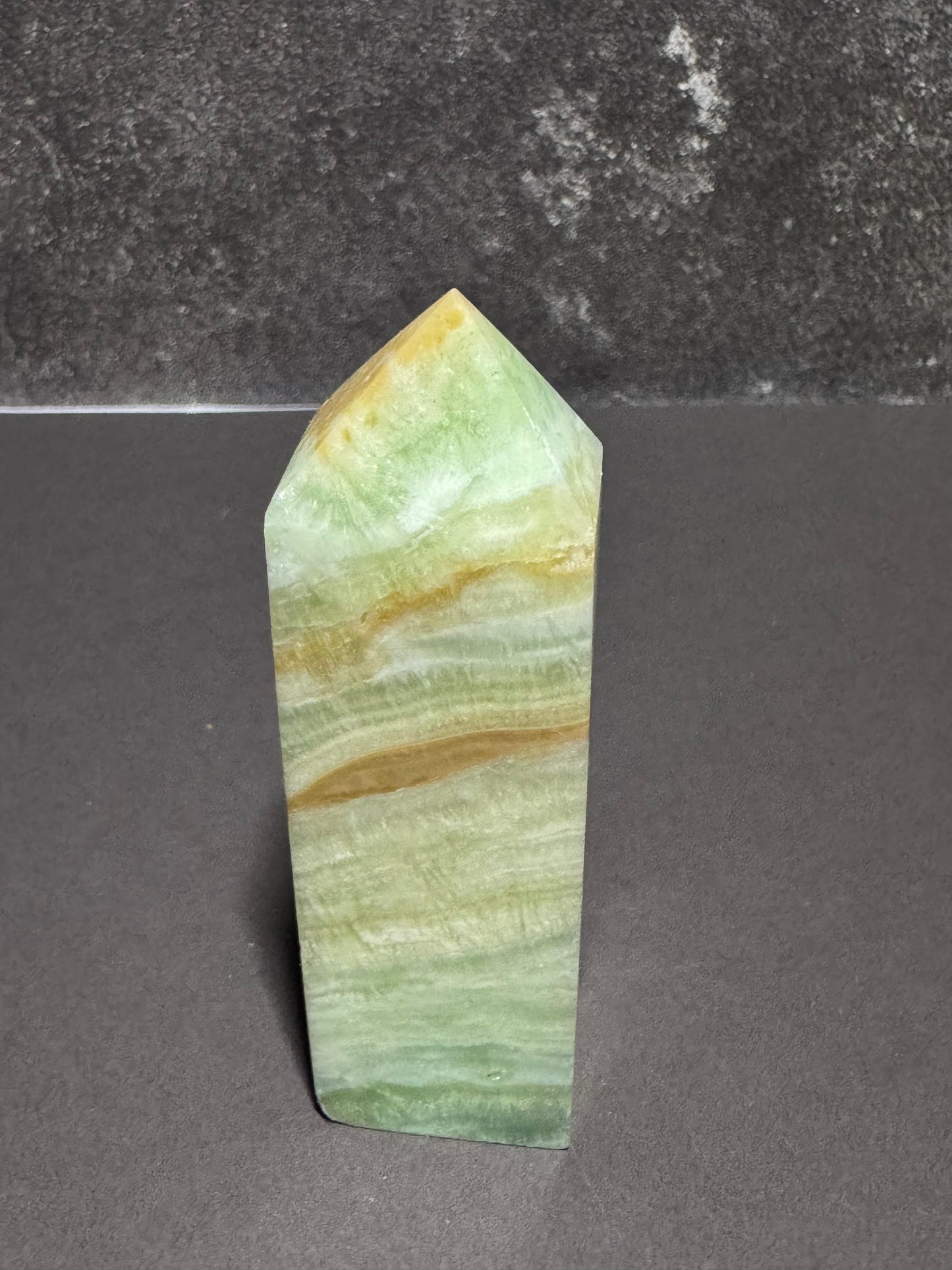 A polished natural crystal point, likely onyx or marble, featuring layered bands of light green, pale yellow, and tan, standing upright on a dark gray surface against a dark gray textured background.
