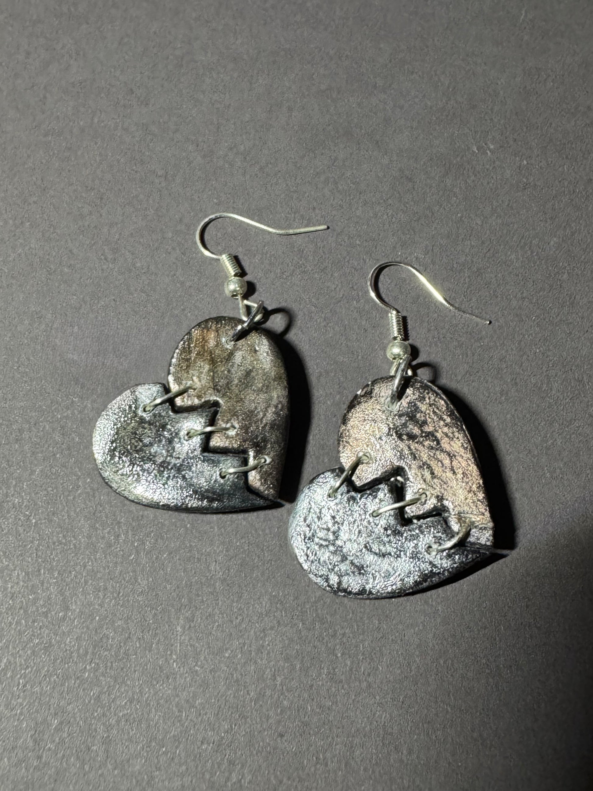 A pair of handmade heart-shaped dangle earrings with a distressed, metallic silver and bronze finish, featuring a broken-heart motif and silver fishhook wires, displayed on a dark gray background.