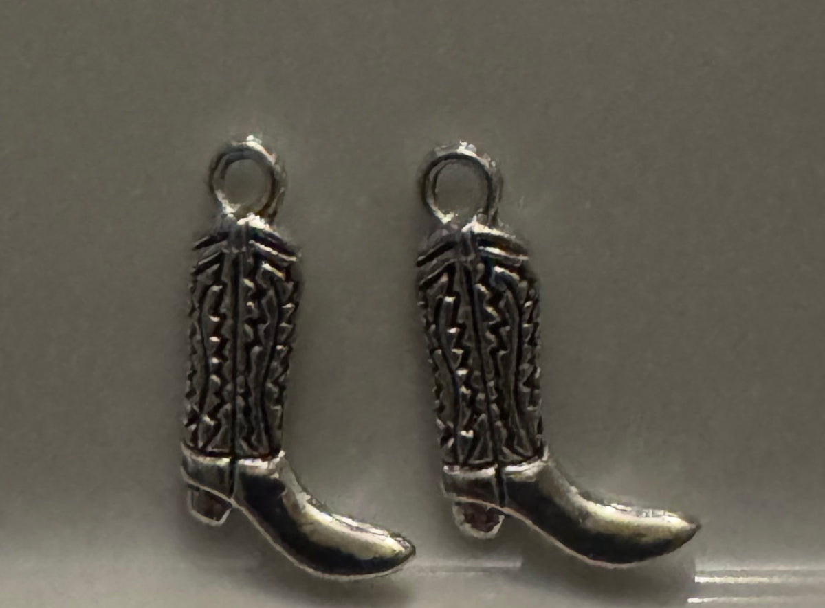 A pair of silver-toned metal cowboy boot charms with detailed engravings and hanging loops, displayed on a neutral gray background.
