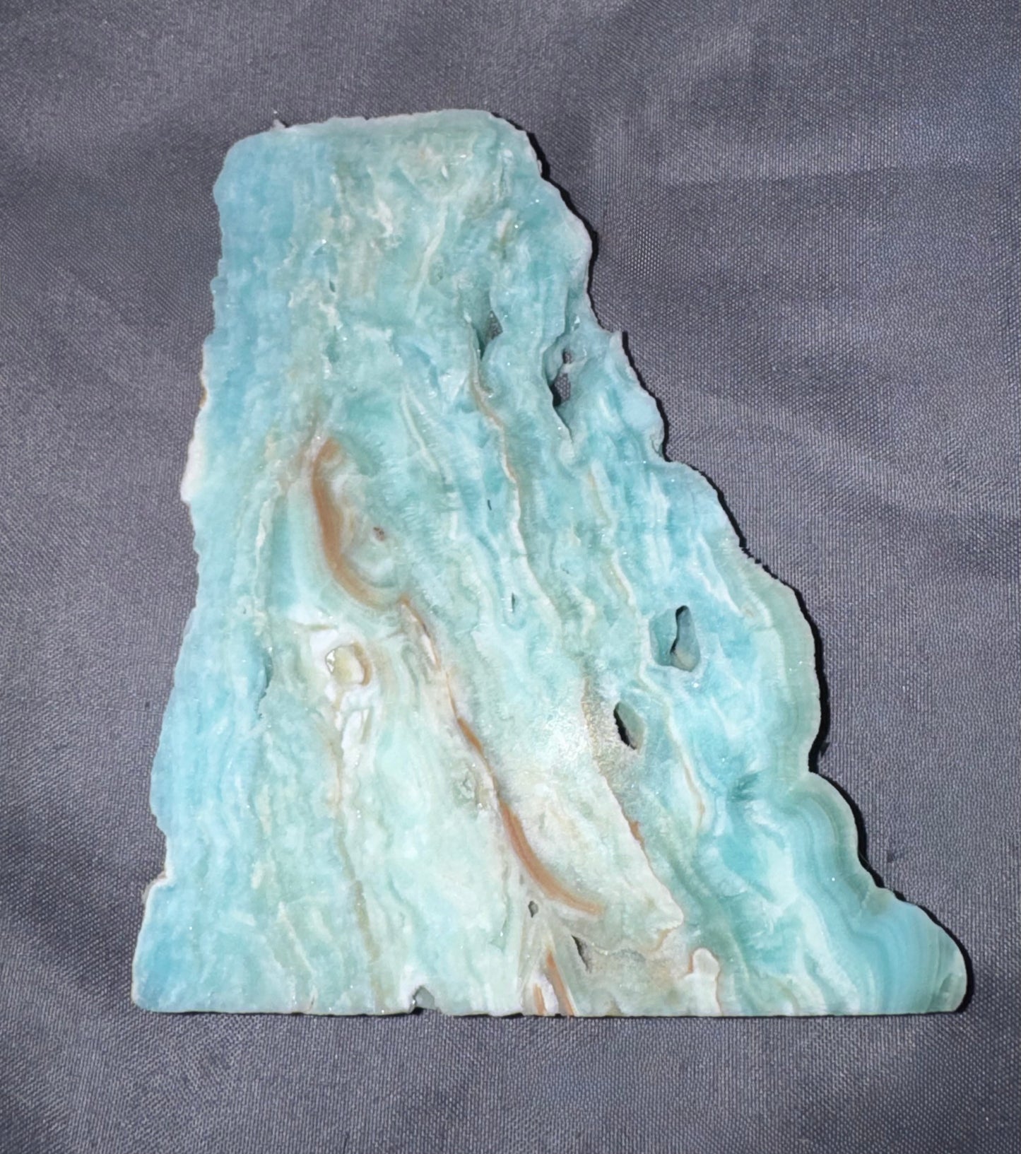 A raw, polished stone slab with natural organic edges, featuring marbled patterns of light blue, turquoise, white, and soft tan veins, displayed on a dark gray textured fabric background.