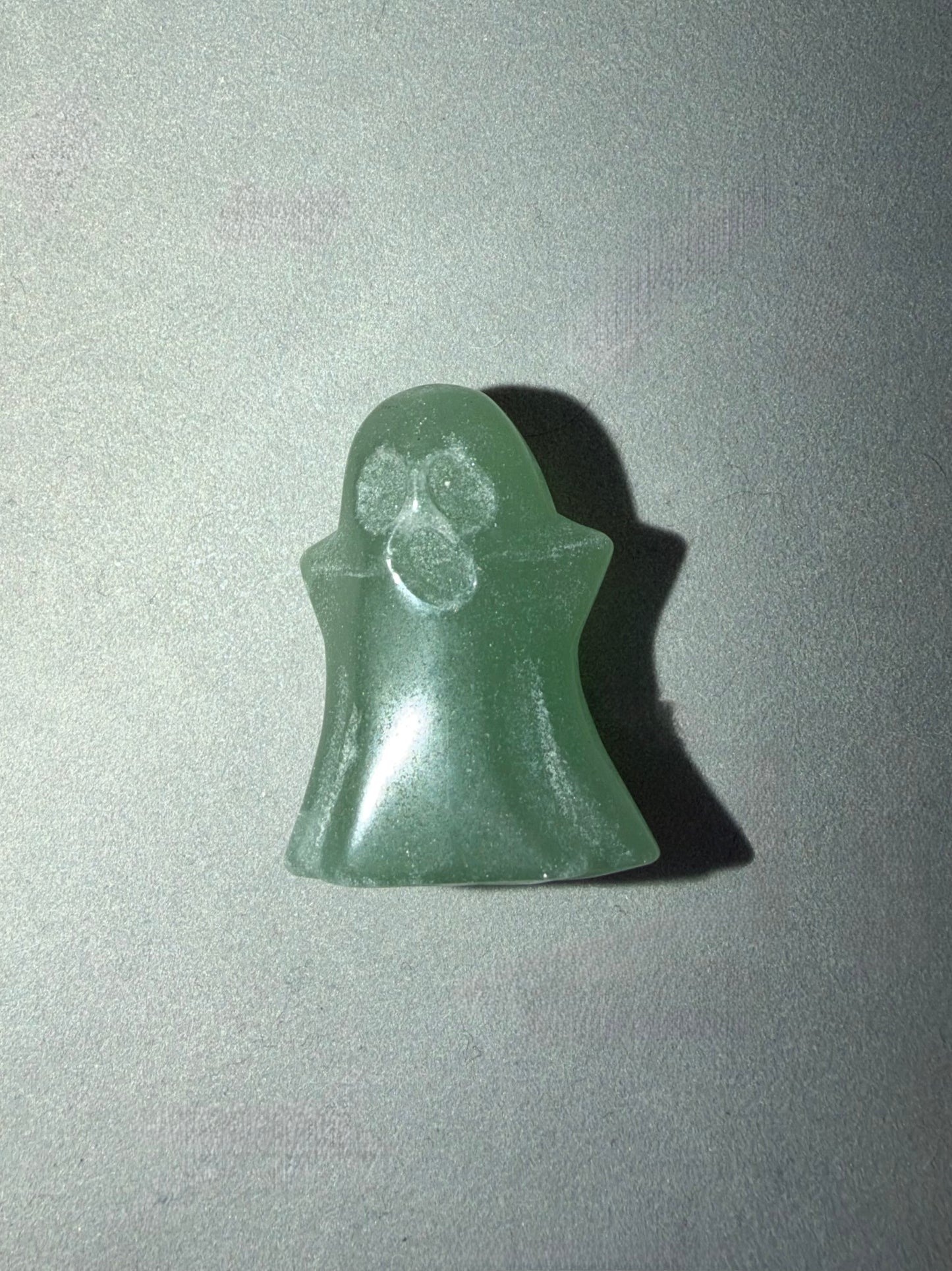 A small ghost-shaped green stone or resin charm with a smooth matte finish, photographed on a light gray background with a hard shadow cast to the right.