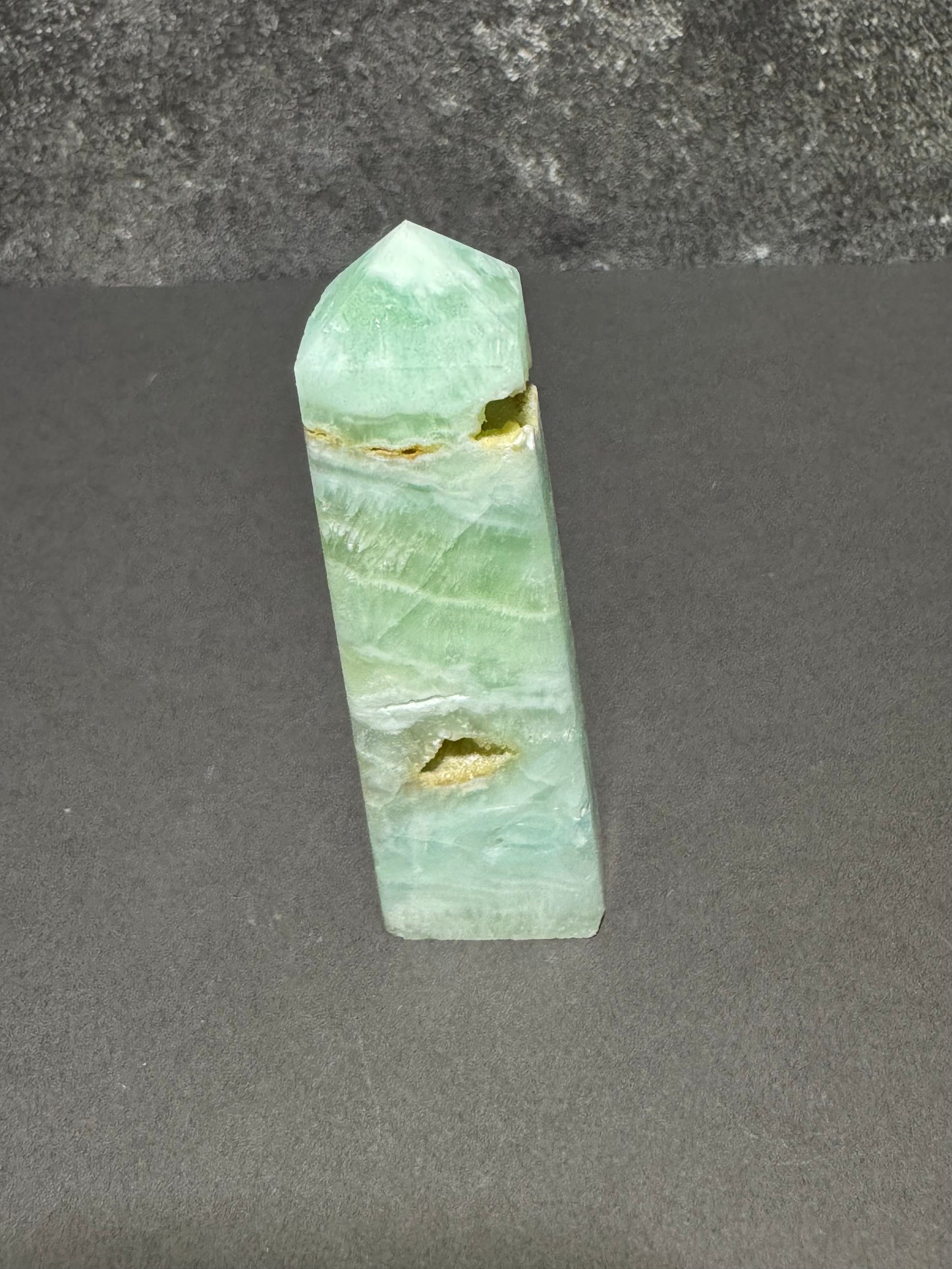 A light green natural crystal point tower, likely fluorite, featuring natural fissures and banded mineral patterns, standing upright on a dark gray surface.