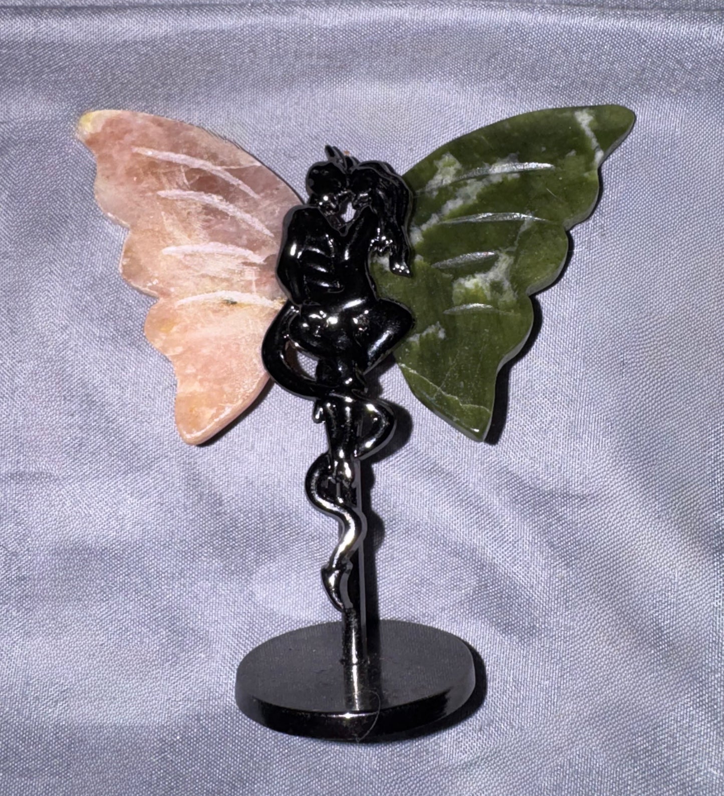 A decorative winged figurine on a black metal stand, featuring a peach-pink stone wing, a dark green stone wing, and a glossy black central figure seated on a snake-like staff over a round black base.